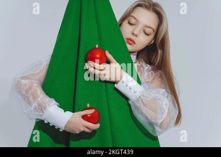 Ein Mädchen mit roten Kugeln, das einen abstrakten Weihnachtsbaum vor weißem Hintergrund umarmt Stockfoto