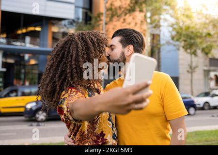 Ein junges Paar küsst sich und macht Selfie über ein Smartphone auf Fußweg Stockfoto