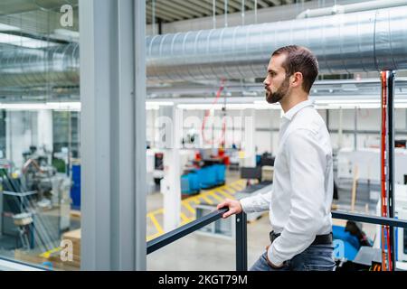 Ein seriöser junger Geschäftsmann mit einer Hand in der Tasche, der in der Fabrik am Geländer steht Stockfoto