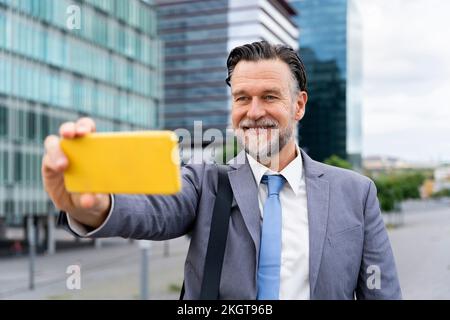 Glücklicher, reifer Geschäftsmann, der Selfie über das Smartphone vor dem Gebäude macht Stockfoto