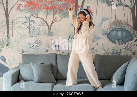 Sorgenfreie junge Frau mit kabellosen Kopfhörern, die zu Hause auf dem Sofa tanzt Stockfoto