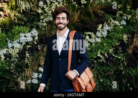 Lächelnder Geschäftsmann mit Schultertasche, der vor Pflanzen steht Stockfoto