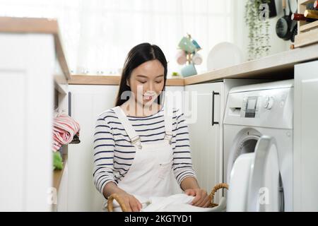 Junge, glückliche Asiatin, die lächelt und zu Hause Wäsche wäscht. Hübsches Mädchen, das Kleider in eine Waschmaschine mit Frontlader legt. Hausarbeit- und Hausarbeit-Konzept Stockfoto