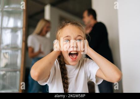 Nahaufnahme des Gesichts einer traurigen kleinen Tochter, die in die Kamera schaut, Ohren verdeckt, während wütende Eltern zu Hause streiten und sich im Hintergrund streiten. Stockfoto