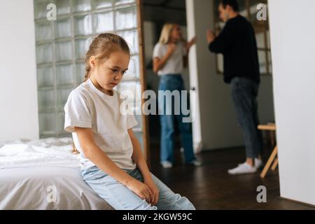 Portrait von deprimierter süßer, kleiner Tochter Traurigkeit, die auf dem Sofa runterschaut, während Eltern sich streiten und im Wohnzimmer im Hintergrund streiten. Stockfoto