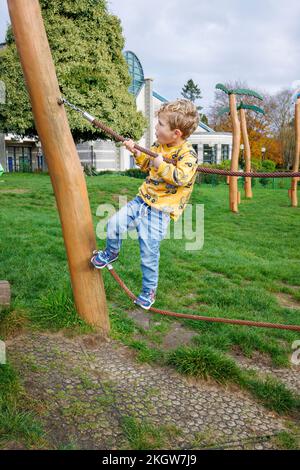 Ein kleiner blonder Kaukasier (im Alter von 4 Jahren) mit gelbem Oberteil und blauer Jeans klettert auf Seile, während er im Woking Park spielt. Wackeln, Surrey Stockfoto