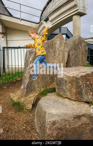 Ein kleiner blonder Kaukasier (im Alter von 4 Jahren), der ein gelbes Oberteil und blaue Jeans trägt, springt beim Spielen im Woking Park von Felsen. Wackeln, Surrey Stockfoto