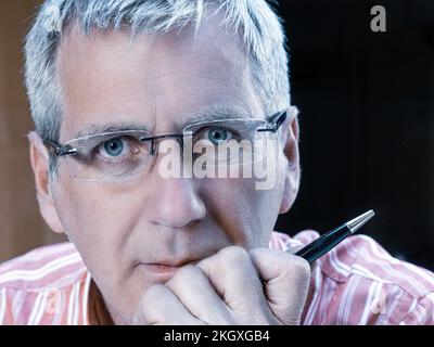 Geschäftsmann männlich 50s-Gesicht, reifer Brillenträger, Hand am Kinn hält einen Stift, der direkt in die Kamera schaut, mit ernstem Gesichtsausdruck Stockfoto