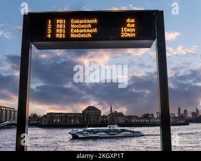 CANARY WHARF CLIPPER RIVER BOAT RB1 Digital Arrivals Information Screen und RB1 Thames Clipper River Boat Arrival at Sunset Arrival in Canary Wharf London UK mit City of London einschließlich The Shard Behind. Stockfoto