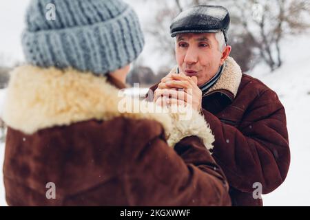 Ein älterer Pflegender wärmt die Hände seiner Frau mit Atem im Freien. Das ältere Familienpaar spaziert im Park bei verschneitem, kaltem Winterwetter. Valentinstag Stockfoto