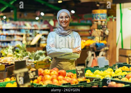 Porträtfrau in einem Hijab als Verkäufer in einem Supermarkt, eine Verkäuferin mit verschränkten Armen lächelt und in die Kamera schaut, eine Schürze verkauft Äpfel und Früchte, eine muslimische Frau ist zufrieden mit Brillen. Stockfoto