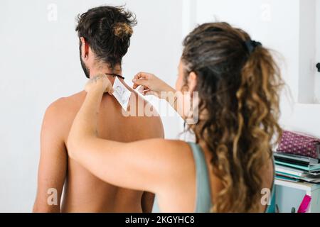 Junge alternative Frau, die einen Tätowierpapierbezug auf den Rücken eines jungen Mannes für die Handstichtechnik legt Stockfoto