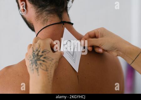 Tätowierte Hände, die einen Tätowierpapierbezug auf den Rücken eines jungen Mannes für die Handstichtechnik legen Stockfoto