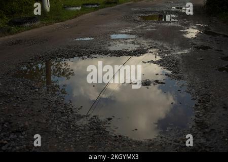 Pfütze auf dem Boden. Grube auf der Straße. Sand und Steine. Reflexion des Himmels in der Pfütze. Stockfoto