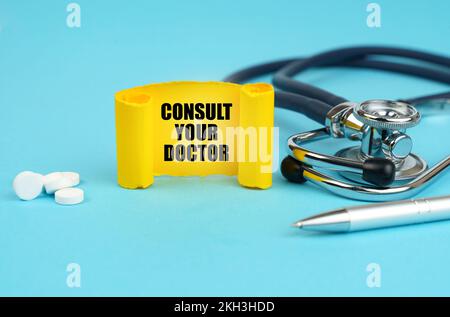 Medizinisches Konzept. Auf einer blauen Oberfläche, einem Stethoskop, Pillen, einem Stift und einem gelben Schild mit der Aufschrift – Wenden Sie sich an Ihren Arzt Stockfoto