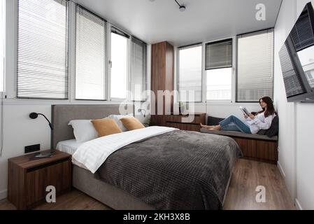 Frau liest Buch in einem Schlafzimmer mit Bett und Holzgarderobe in einem modernen Apartment Stockfoto