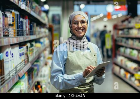 Porträt einer Frau in einem Hijab, einer Ladenarbeiterin in einem Supermarkt mit einem Tablet-Computer, schaut in die Kamera und lächelt, eine Verkäuferin in Gläsern zwischen Regalen mit Waren. Stockfoto