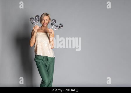 Frohe Neujahrsfeier 2023. Frau mit goldenen Ballons in Form von Zahlen auf grauem Hintergrund. . Hochwertiges Foto Stockfoto