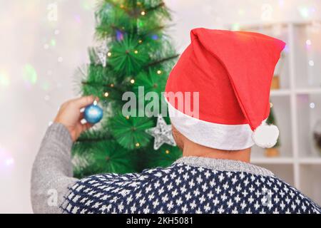 Vorbereitung auf das neue Jahr. Einrichtung zu Neujahr und Weihnachten. Ein Mann schmückt einen Baum mit Spielzeug. Vorbereitung auf Weihnachten. Ein Mann in einem Weihnachtshut schmückt einen Weihnachtsbaum für den Feiertag. Stockfoto