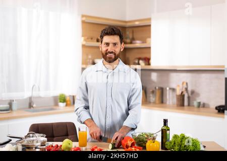 Lächelnder, gutaussehender, erwachsener, bärtiger europäischer Koch, der Bio-Gemüse für Salat schneidet Stockfoto