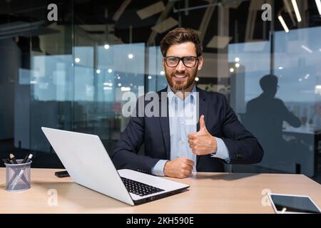 Porträt eines reifen erfolgreichen Investors, eines Geschäftsmanns im Geschäftsanzug, der im Büro mit einem Laptop arbeitet, eines Senior-Chefs, der die Kamera lächelt und Daumen nach oben zeigt. Stockfoto