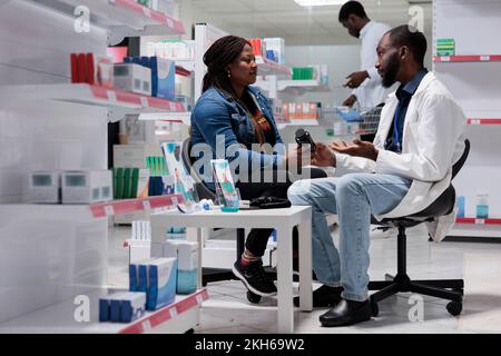 Apothekenberatung, Patient kauft Pillen Rezept, Apotheker gibt Vitaminflasche, Seitenansicht. Ein Kunde nimmt Medikamente, ein Chemiker und ein Kunde spricht, alles schwarzes Team Stockfoto