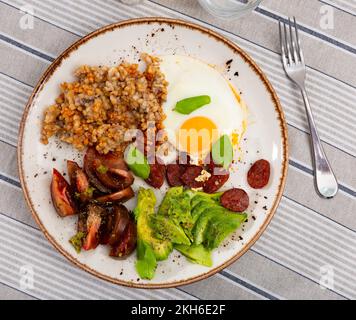 Köstliches Frühstück mit Buchweizen, sonnengetrockneten Tomaten, Avocado, Spiegeleiern mit Basilikum und Jagdwürsten Stockfoto