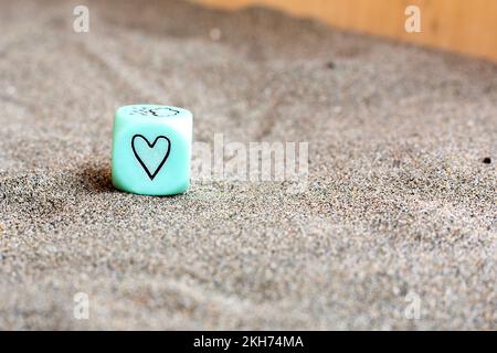 hellblauer Kunststoffwürfel mit Herz- und Wettersymbolen an den Seiten. Lustige Spielwürfel auf Sand. Valentinstag-Konzept. Liebessymbol. Schließen Stockfoto