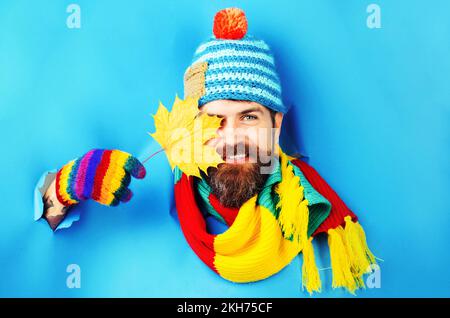 Lächelnder bärtiger Mann in Strickmütze, Schal und Handschuhen bedeckte das Auge mit Ahornblatt im Herbst. Stockfoto