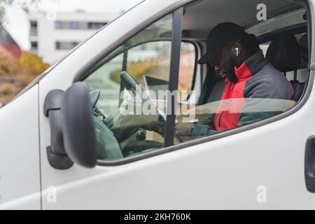 Technologie, die bei der Paketzustellung verwendet wird. Außenansicht eines professionell fokussierten afroamerikanischen Post-Delivery-Mannes, der im weißen Lieferwagen auf dem Fahrersitz sitzt und seine Checkliste mit einem weißen, kabellosen Ohrhörer im Ohr ansieht. Hochwertiges Foto Stockfoto