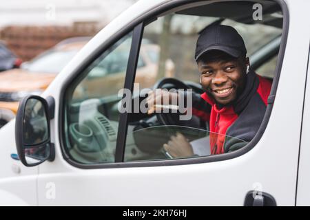 Junger erwachsener schwarzer Liefermann sitzt auf dem Fahrersitz eines weißen Lieferwagens und lehnt sich lächelnd aus dem Fenster. Lieferservice. Horizontale Aufnahme. Hochwertiges Foto Stockfoto