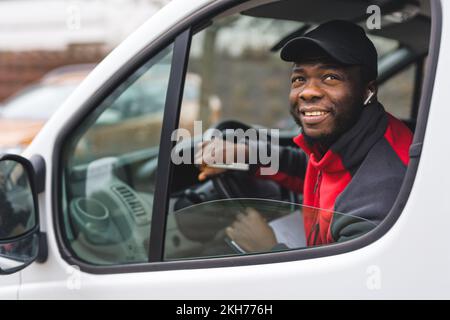 Positives Außenporträt eines freundlichen, gutaussehenden Schwarzen Mannes mit schwarzem Hut und roter Jacke, der als Paketkurier arbeitet. Im Fenster Lieferwagen anzeigen. Hochwertiges Foto Stockfoto