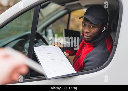 Verschwommene Hand mit einem Stift eines nicht erkennbaren Kaukasiers, der auf Lieferdokumente zusteuert, die auf dem Klemmbrett des gutaussehenden afroamerikanischen Lieferers in schwarzem Baseballhut stehen. Hochwertiges Foto Stockfoto