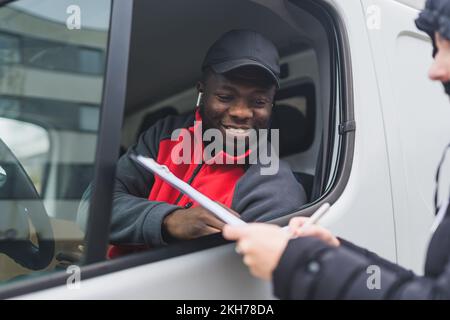 Positive menschliche Interaktionen. Freundlicher afroamerikanischer Kurier in rot-schwarzer Uniform, der einem nicht wiedererkennbaren Kaukasier beim Ausfüllen der Lieferpapiere hilft. Transportkonzept. Hochwertiges Foto Stockfoto