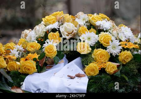 Grabblumen mit gelben Rosen und weißer Trauerschleife auf einem Grab mit herabfallenden Herbstblättern Stockfoto