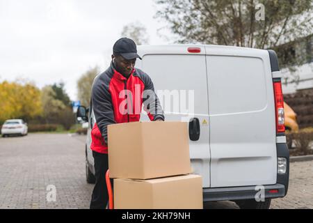 Paketzustellkonzept. Schwarzer, gutaussehender männlicher Kurier, der große Pakete in Kartons mit einem Handwagen ausliefert. Weißer Lieferwagen im Hintergrund. Hochwertiges Foto Stockfoto