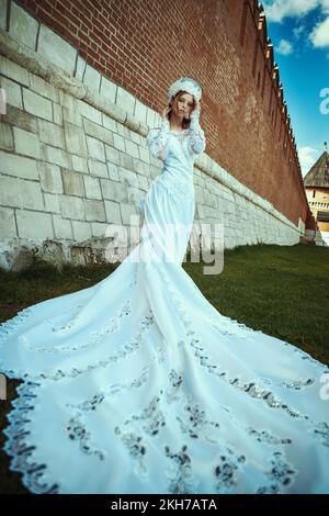 Ein Mädchen in einem langen Hochzeitskleid und einem Kokoshnik in der Nähe einer alten Mauer. russische zaritsa Stockfoto