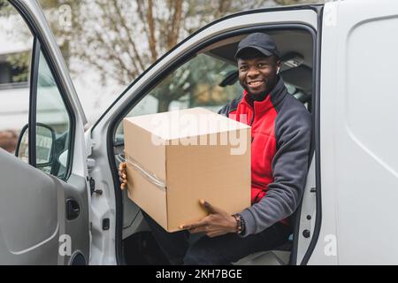 Freundlicher, lächelnder Schwarzer in der Mitte der 20 Jahre, der auf dem Fahrersitz sitzt, die Tür geöffnet hat und ein Papppaket in der Hand hat. Ich schaue in die Kamera. Positive Schwingungen. Hochwertiges Foto Stockfoto