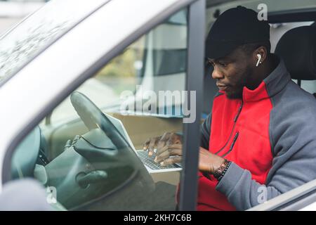 Ein müder, aber fokussierter afroamerikanischer Liefermann, der an seinem Laptop arbeitet, während er auf dem Fahrersitz sitzt. Durch das Fahrerfenster geschossen. Versand und Lieferung. Hochwertiges Foto Stockfoto