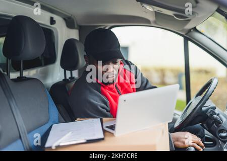 Fahrzeuginterne Ansicht eines fokussierten schwarzen Zustellers mittleren Alters in Uniform, der sein silbernes Notebook zum Durchstreichen der nächsten Pakete verwendet. Die Nutzung des Fahrzeugs - weißer Lieferwagen. Hochwertiges Foto Stockfoto