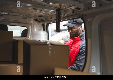 Junger erwachsener schwarzer Liefermann mit rotem Uniformpullover, der in einen Lieferwagen voller Papppakete schaut. Horizontale Aufnahme. Hochwertiges Foto Stockfoto