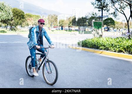 Ein Latino in Jeans, der Fahrrad fährt, in einer Stadt, mit roter Mütze und Brille. Konzept des ökologischen Transports Stockfoto