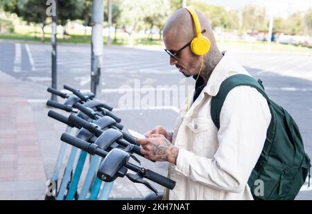 Kahler junger Mann mit gelben Kopfhörern, der eine Handy-App benutzt, um einen öffentlichen Roller in der Stadt zu fahren Stockfoto
