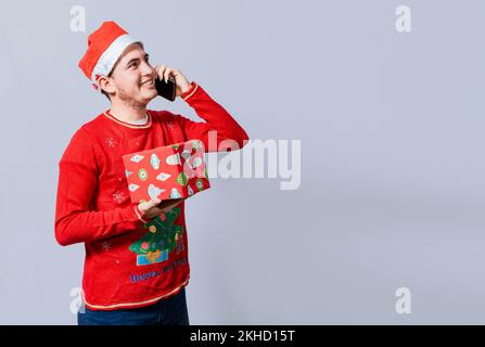Ein Weihnachtsmann, der ein Geschenk hält und seine Familie telefoniert. Glücklicher Mann, der ein Geschenk hält und Familie zu weihnachten anruft, lächelnder Kerl mit weihnachtsmütze, der eine C hält Stockfoto