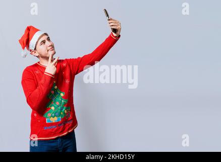 Ein glücklicher gutaussehender Mann mit weihnachtsmütze, der ein Selfie macht. Ein glücklicher Kerl, der ein weihnachtsselfie auf isoliertem Hintergrund macht, lächelnder junger Mann in einem weihnachtshut Stockfoto