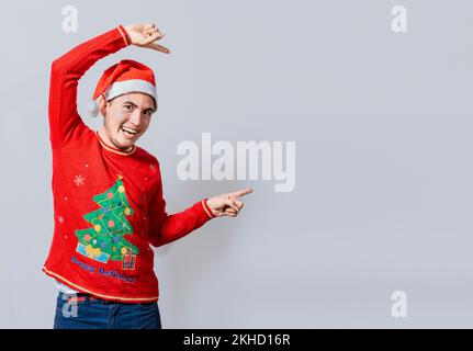 Freundlicher Weihnachtsmann, der auf einen Platz zur Seite zeigte. Freundlicher, lächelnder junger Mann in weihnachtskleidung, der auf eine Werbeaktion zeigt, fröhlicher weihnachtsmann CO Stockfoto