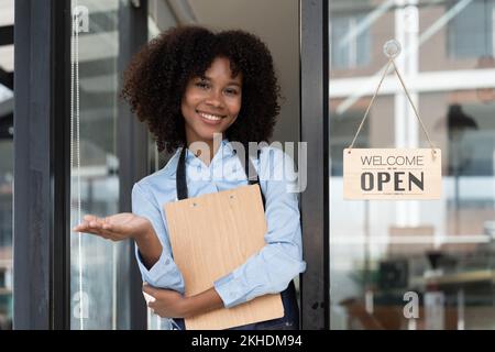 Kleine afrikanische Eigentümerin lächelt, während sie das Schild für die Eröffnung des Cafés umdreht. Fröhliche afro-amerikanische Kellnerin Unternehmerin in Schürze Geschenk Stockfoto