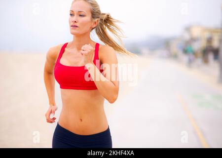 Sie treibt sich bei jedem Lauf weiter. Eine junge Frau, die am Strand joggt. Stockfoto