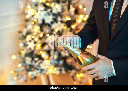 Nahaufnahme eines Mannes im Anzug, der eine Flasche Champagner öffnet, da ist ein weihnachtsbaum im Hintergrund. Stockfoto