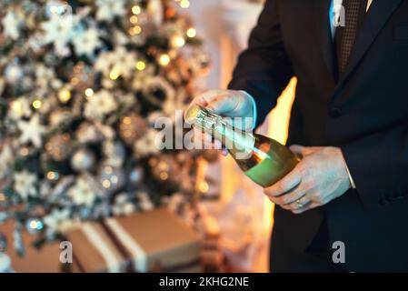 Nahaufnahme eines Mannes im Anzug, der eine Flasche Champagner öffnet, da ist ein weihnachtsbaum im Hintergrund. Stockfoto